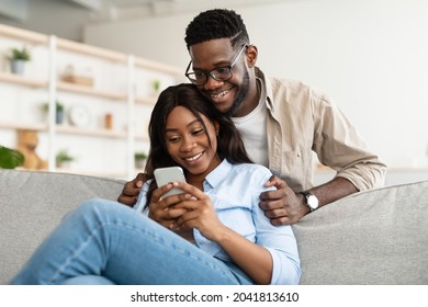 Smiling young black man in braces embracing woman from behind while looking at smartphone. African American couple sharing social media on cellphone, happy female sitting on the sofa, showing gadget - Powered by Shutterstock