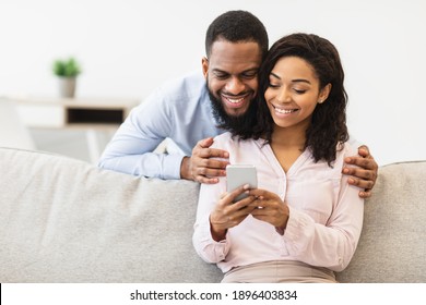 Smiling Young Black Man Embracing Woman From Behind While Looking At Smartphone. African American Couple Sharing Social Media On Mobile Phone, Happy Female Sitting On The Couch, Showing Gadget