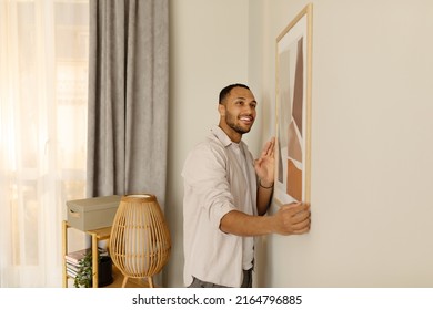 Smiling young black man putting picture frame, hanging painting on wall, empty space. African American male interior designer decorating new modern stylish apartment. Home interior and domestic decor - Shutterstock ID 2164796885