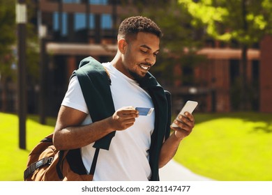 Smiling Young Black Man Using Smartphone And Credit Card In Park, Online Shopping Concept