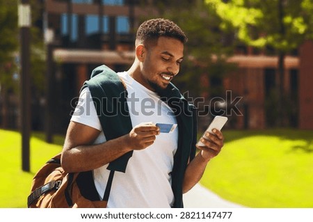 Smiling Young Black Man Using Smartphone And Credit Card In Park, Online Shopping Concept