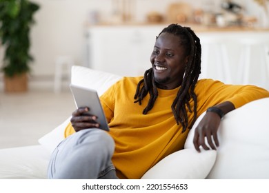 Smiling Young Black Man Watching Videos On Digital Tablet While Resting On Couch In Living Room, Happy Millennial African American Guy Relaxing At Home With Modern Gadget, Enjoying Weekend Pastime