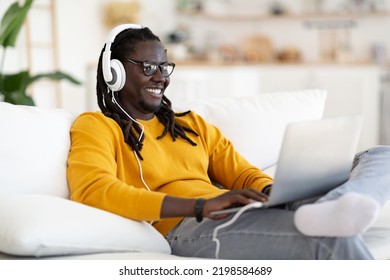 Smiling Young Black Man Wearing Wireless Headphones Relaxing With Laptop At Home, Happy African American Man In Eyeglasses Sitting On Comfortable Couch In Living Room And Using Computer, Copy Space
