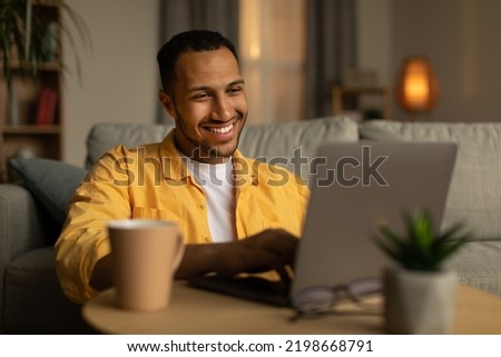 Smiling young black man working or communicating online, sitting on couch with laptop at home. Cheerful millennial guy freelancing, chatting on internet. Modern technologies in everyday life concept