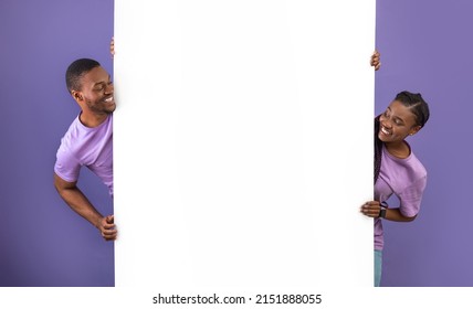 Smiling young black people holding white vertical advertisement board, demonstrating free copy space for your text or design, positive guy and lady peeking out banner, purple violet background wall