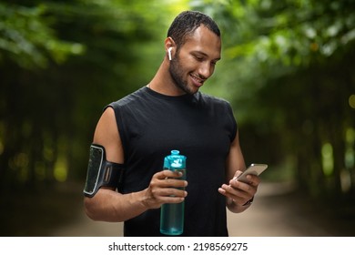 Smiling young black sportsman exercising at public park, using smartphone and wireless earbuds, checking mobile fitness application, drinking water while having break, copy space - Powered by Shutterstock