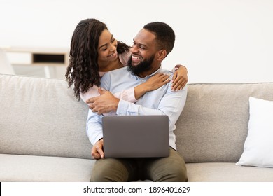 Smiling Young Black Woman Embracing Man From Behind While He Using Laptop. Loving African American Couple Spending Time Together, Happy Male Sitting On The Couch Working On Computer