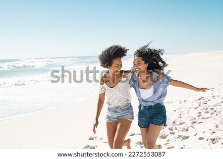 Smiling young black woman having fun at beach with her best friend. Latin hispanic young women running on seashore barefoot during vacation. Cheerful friends enjoying at sea on a bright sunny day.