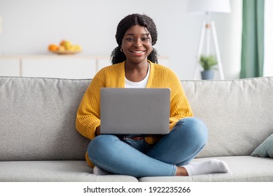 Smiling Young Black Woman In Knitted Sweater Working From Home, Sitting On Couch And Using Laptop, Copy Space. Relaxed African American Lady Enjoying Her Weekend, Surfing On Internet On Laptop