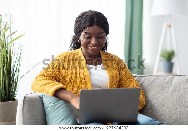 Smiling young black woman in knitted sweater\
freelancer working from home, sitting on couch, using laptop, copy\
space. Relaxed african american lady enjoying her weekend, surfing\
on internet on laptop