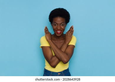 Smiling Young Black Woman With Short Afro Hair Is Showing Letter X With Arms Crossed. Front View. Waist Up Studio Portrait On Blue Background.