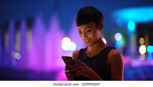 Smiling Young Black Woman Texting On Cell Phone Colorful Fountain In Background