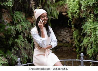 Smiling young black woman wearing a beige wool hat sits on a railing in a lush garden, engaged in a conversation on her smartphone while enjoying the serene surroundings - Powered by Shutterstock