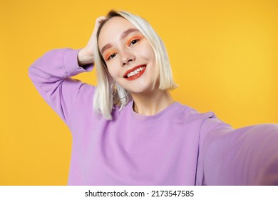 Smiling young blonde caucasian woman bob haircut bright makeup wearing basic purple shirt close up doing selfie shot on mobile phone open mouth isolated on yellow color background studio portrait