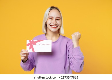 Smiling young blonde caucasian woman bob haircut bright makeup wearing casual basic purple shirt hold gift voucher doing winner gesture clenching fists isolated on yellow background studio portrait