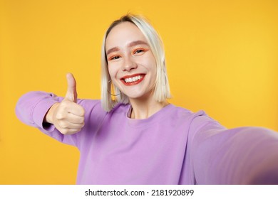 Smiling young blonde caucasian woman bob haircut bright makeup wears basic purple shirt close up doing selfie shot on mobile phone showing thumbs up isolated on yellow color background studio portrait