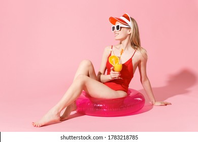 Smiling Young Blonde Girl In Red One-piece Swimsuit Cap Sunglasses Isolated On Pink Background. People Summer Vacation Rest Lifestyle Concept. Sitting In Swim Inflatable Ring Hold Glass With Cocktail