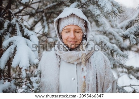 Smiling young blonde girl stands with closed eyes under snowfall in winter forest