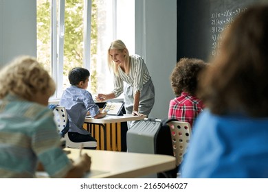 Smiling Young Blonde Teacher Tutor Explains Lesson To Schoolboy Using Tablet Device Teaching Mathematics Elementary Middle Diverse Schoolchildren Standing At Chalkboard Blackboard. Education Concept.