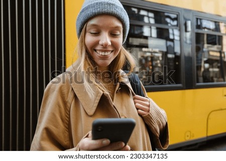 Smiling young blonde woman casually dressed using smartphone while standing at bus station