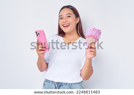 Smiling young blonde woman girl Asian wearing casual white t-shirt using mobile phone and holding money rupiah banknotes isolated on white background. Fast credits concept