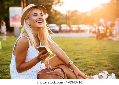 Smiling Young Blonde Woman Listening To Music While Sitting On Grass In Park