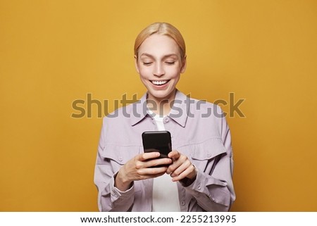 Smiling young blonde woman posing isolated on yellow studio wall background. People lifestyle concept. Model using mobile phone, typing sms message