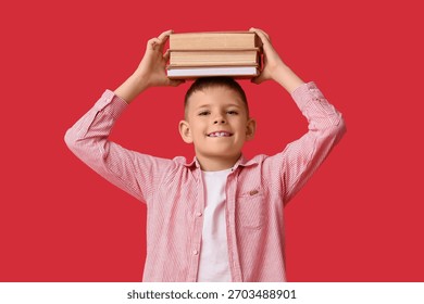Smiling young boy balancing stack of books on head, education and learning concept, childhood development, red background