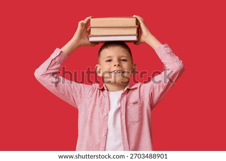 Smiling young boy balancing stack of books on head, education and learning concept, childhood development, red background