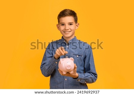 Smiling young boy in blue shirt depositing coins into pink piggy bank, happy teen male child illustrating financial responsibility and savings concept, standing isolated against yellow background
