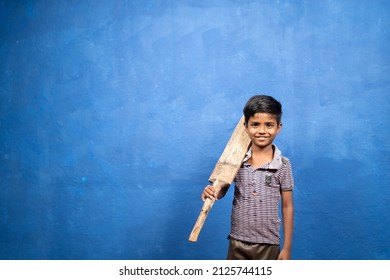 Smiling young boy kid holding cricket bat by looking at camera - concept of future goal, inspiration, childhood dream and poverty