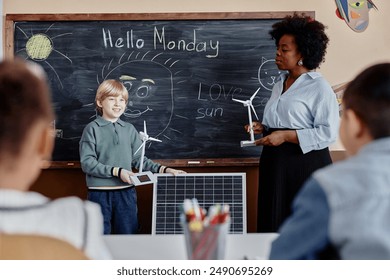 Smiling young boy of primary school showing big solar panel and small wind turbine to classmates answering at blackboard with funny sun drawings - Powered by Shutterstock