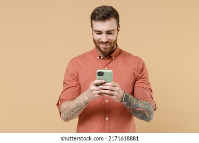 Smiling young brunet man 20s he wears orange shirt hold in hand use mobile cell phone chat on mobile cell phone isolated on plain pastel light beige background studio portrait. Tattoo translate fun