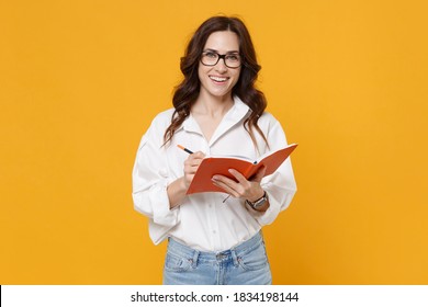 Smiling Young Brunette Business Woman In White Shirt Glasses Isolated On Yellow Background Studio Portrait. Achievement Career Wealth Business Concept. Mock Up Copy Space. Writing Notes In Notebook