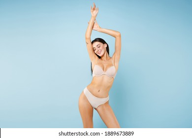 Smiling young brunette woman 20s in beige underwear showing fit body standing with hands up showing result on underarms removing hair procedure isolated on pastel blue wall background studio portrait