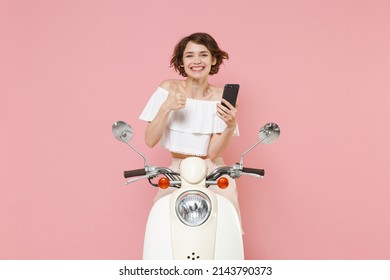 Smiling young brunette woman 20s wearing white summer clothes using mobile cell phone typing sms message showing thumb up sit driving moped isolated on pastel pink colour background studio portrait