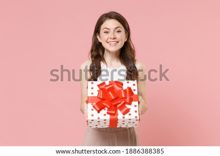 Smiling young brunette woman 20s in light casual clothes posing isolated on pastel pink wall background. Birthday holiday concept. Mock up copy space. Hold white red present box with gift ribbon bow