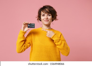 Smiling Young Brunette Woman Girl In Yellow Sweater Posing Isolated On Pastel Pink Wall Background Studio Portait. People Lifestyle Concept. Mock Up Copy Space. Point Index Finger On Credit Bank Card