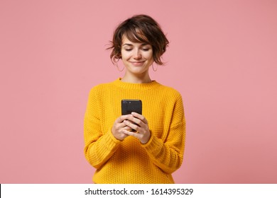 Smiling young brunette woman girl in yellow sweater posing isolated on pastel pink wall background studio portait. People lifestyle concept. Mock up copy space. Using mobile phone, typing sms message