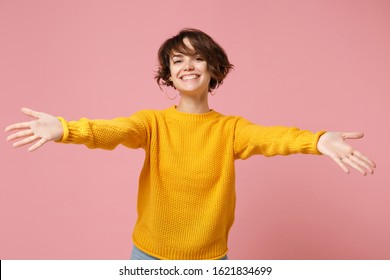 Smiling young brunette woman girl in yellow sweater posing isolated on pastel pink wall background studio portrait. People lifestyle concept. Mock up copy space. Standing with outstretched hands