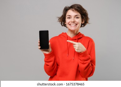 Smiling young brunette woman girl in casual red hoodie posing isolated on grey background. People lifestyle concept. Mock up copy space. Pointing index finger on mobile phone with blank empty screen - Powered by Shutterstock