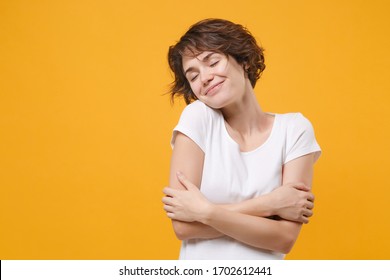 Smiling young brunette woman girl in white t-shirt posing isolated on yellow orange background. People lifestyle concept. Mock up copy space. Keeping eyes closed holding hands crossed hugging herself