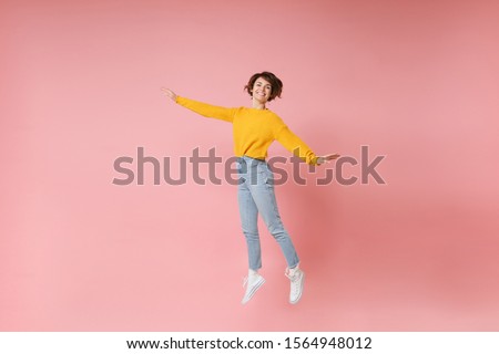 Smiling young brunette woman girl in yellow sweater posing isolated on pastel pink background in studio. People lifestyle concept. Mock up copy space. Having fun spreading hands rising hands, jumping