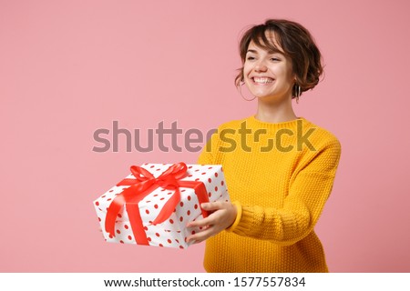 Smiling young brunette woman girl in yellow sweater posing isolated on pink background. Valentine's Day Women's Day birthday holiday concept. Mock up copy space. Hold present box with gift ribbon bow