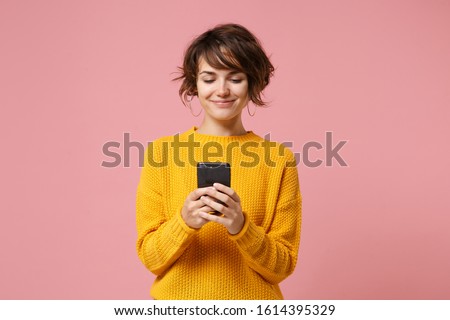 Smiling young brunette woman girl in yellow sweater posing isolated on pastel pink wall background studio portait. People lifestyle concept. Mock up copy space. Using mobile phone, typing sms message