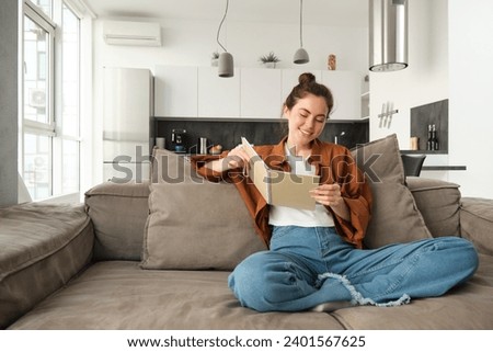 Smiling young brunette woman, sits on sofa in living room, holds notebook, reads her notes, studies for exam, student does her homework at home.