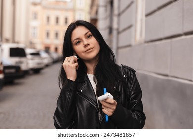 Smiling young brunette woman walking outdoors and putting wireless earphones on. Woman is relaxing listening music in town. Young woman using technologies, wear black leather jacket, hold case. - Powered by Shutterstock