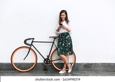 Smiling young brunette woman wearing summer clothes standing at the wall outdoors, holding bicycle, using mobile phone - Powered by Shutterstock