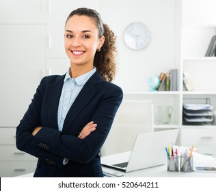 Smiling young business lady with dark hair standing near office desk 