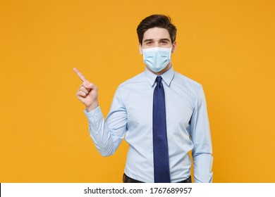 Smiling Young Business Man In Blue Shirt Tie Sterile Face Mask Isolated On Yellow Background. Epidemic Pandemic Coronavirus 2019-ncov Sars Covid-19 Flu Virus Concept. Pointing Index Finger Aside Up